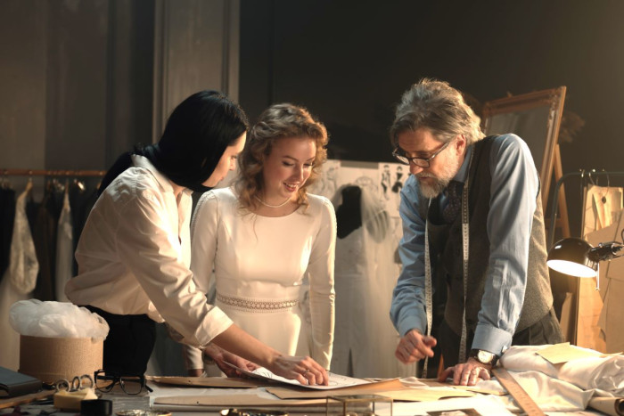 A woman in modern dress looks at plans with an actor in a period costume and a tailor 