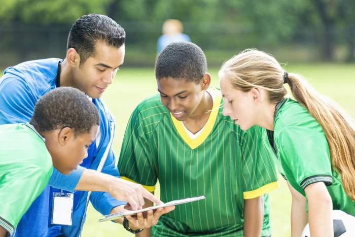 A man shows a clip board to a group of 1 female and 2 male teenagers in sports uniform 